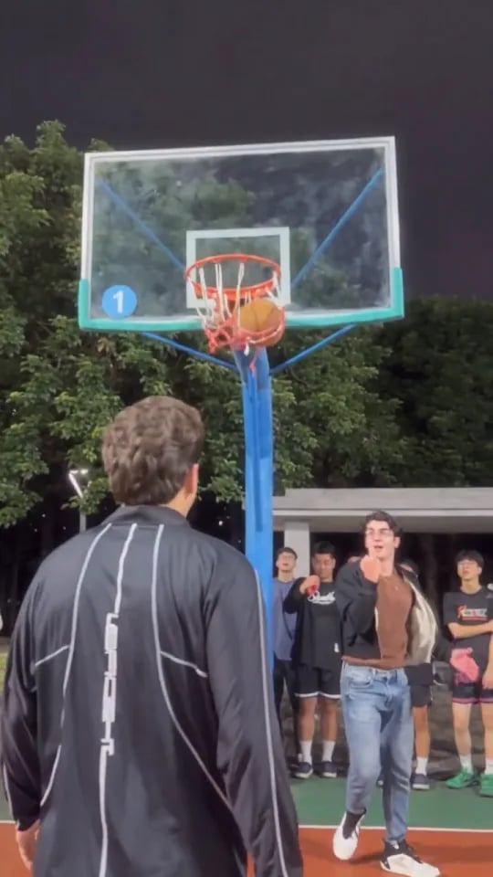The defender in a black long-sleeve shirt, seen from behind, faces a player in a brown sweater on a floodlit court with a crowd behind