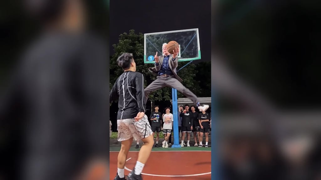 An elderly man in a grey suit leaps past the defender and dunks one-handed on a night court as the crowd watches