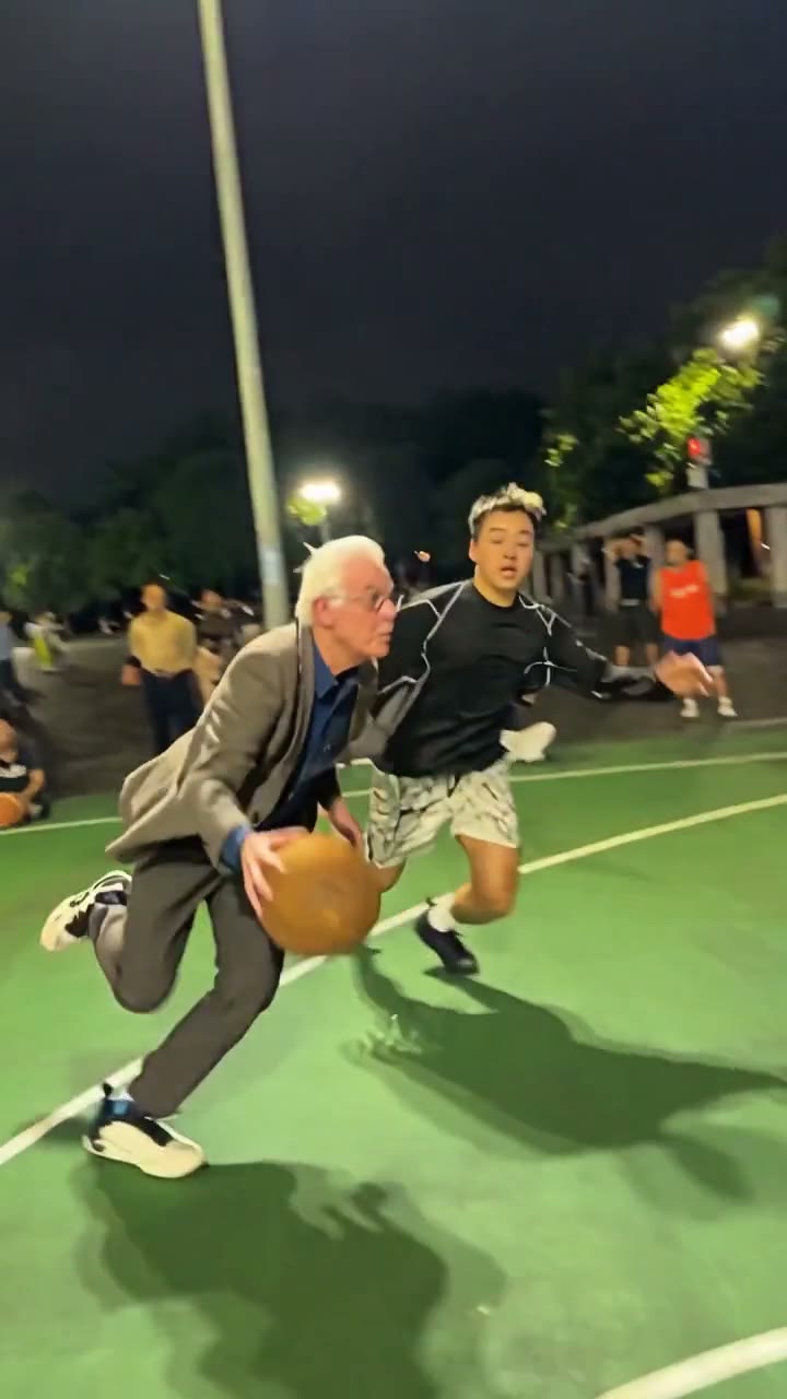 An elderly man in a grey suit drives past the defender with the ball on a night court, with a crowd along the sideline